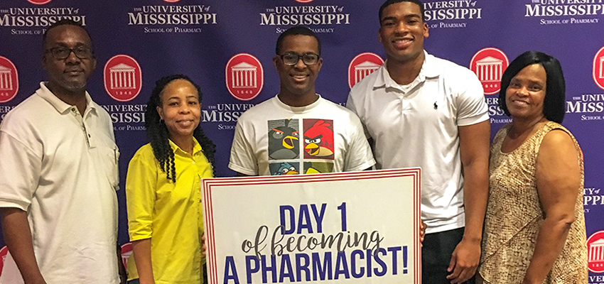 A male student holds a "Day 1 of Becoming Pharmacist" sign while standing with his family.