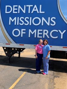 Picture of two females standing outside in front of a semi-truck trailer that reads "Dental Mission of Mercy"