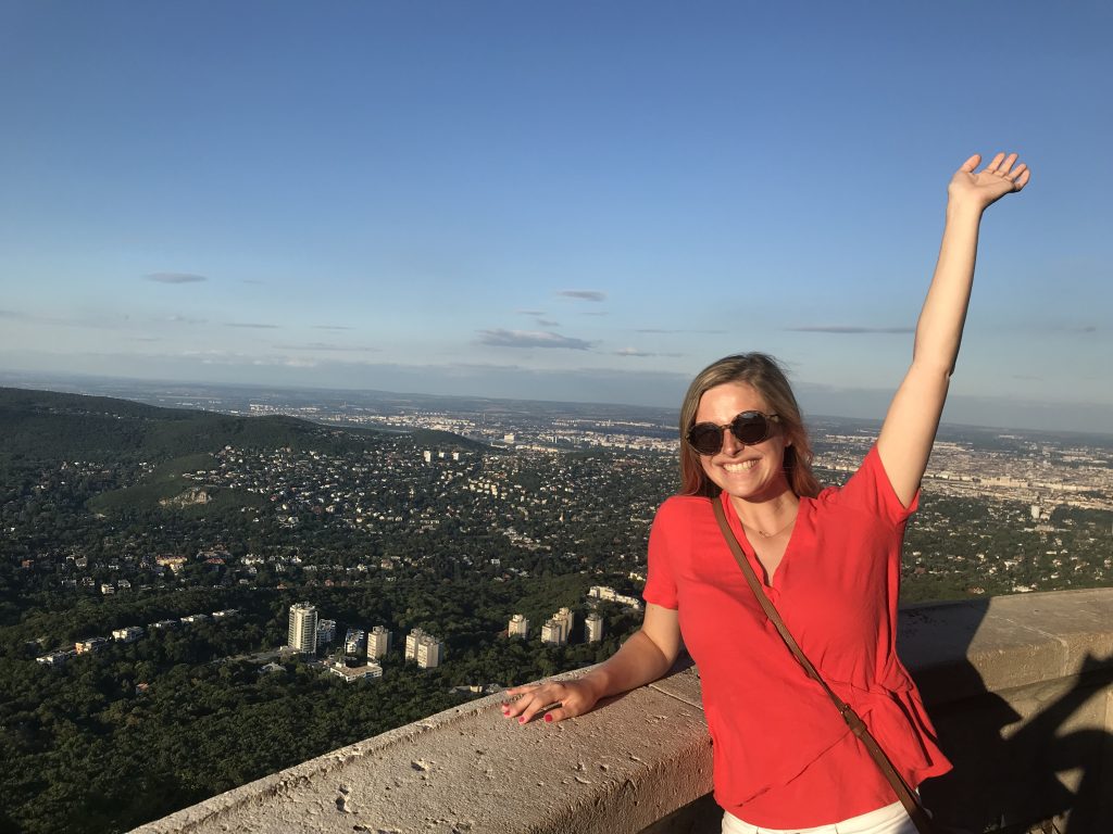 Sophie smiles with hand in the air with city view behind her.