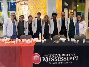 Nine student pharmacists in their white coats stand in a row, smiling