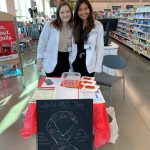 Two female students in white coats standing behind a table located in drug store.