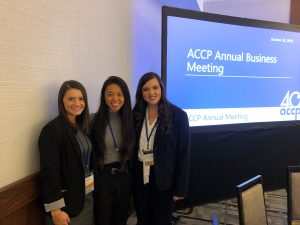 Three female students smile for the camera with screen in background reading "ACCP Annual Business Meeting"