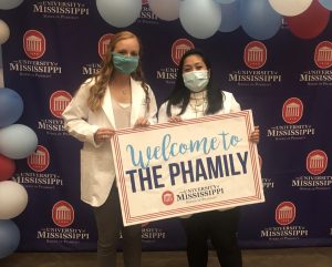 Two female students in white coats and masks holding a sign "Welcome to the Phamily"