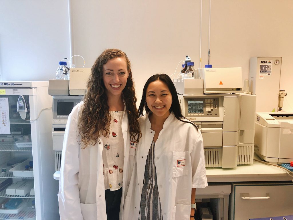 Two female students wear lab coats with research equipment behind them.