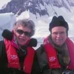 Raye and David Newton wearing cold-weather gear with mountains behind them in Antartica.