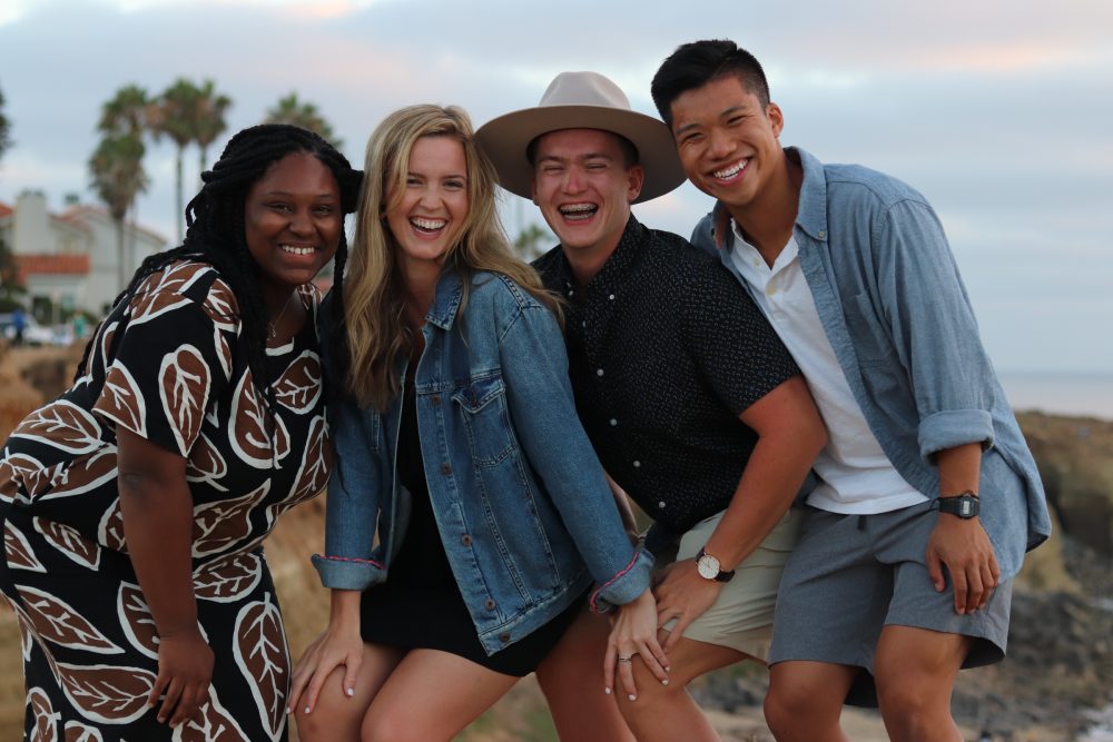 Four students stand together with scenic sunset beach background.