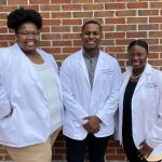 Three student pharmacists stand together wearing their white coats.