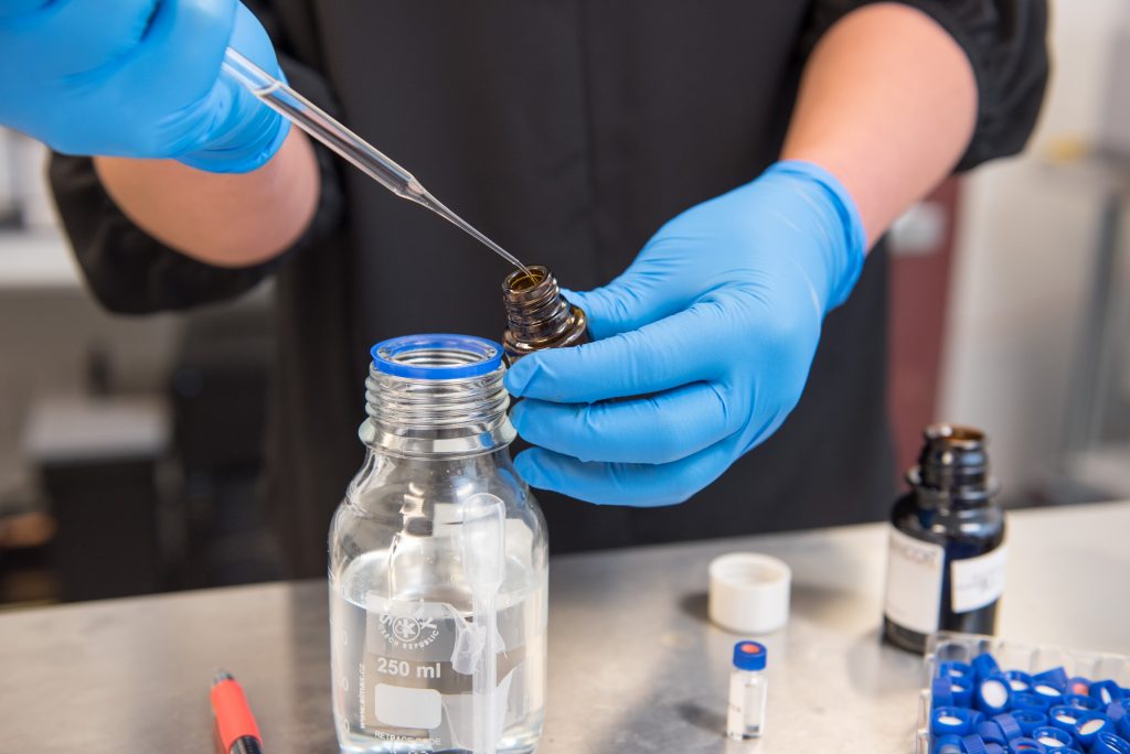 A researcher wears blue gloves as they use a droplet to extract and place oil in glass jars.