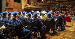 Graduates in cap and gown sit on stage looking out to audience.